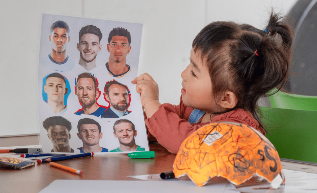 A young girl points at a sheet featuring football players while sitting beside a partially decorated football.